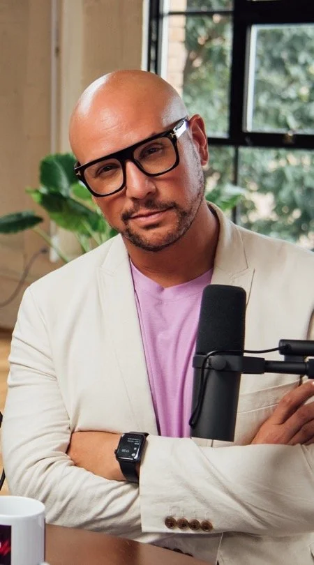 Dr. James Q. Simmons wearing glasses and a blazer, seated behind a podcast microphone in front of a window and indoor plant