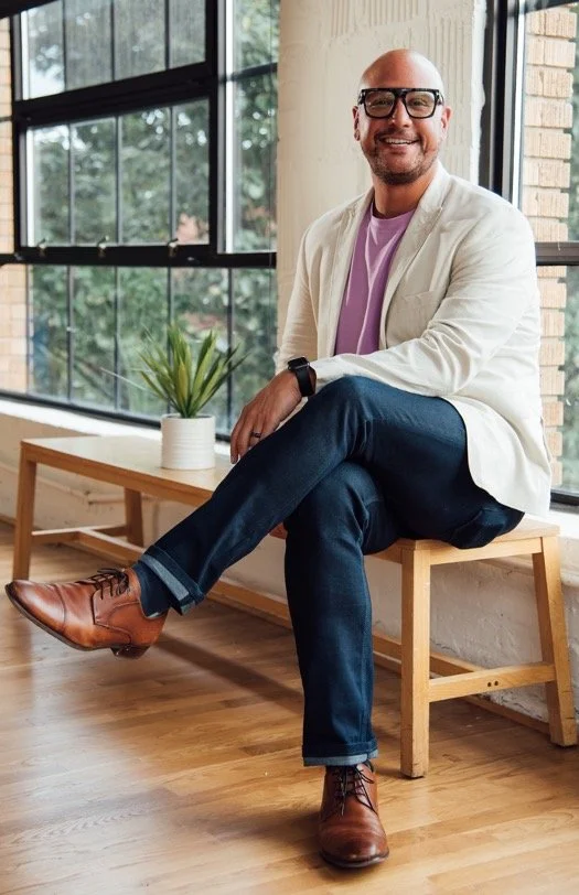 Dr. James Q. Simmons seated in a modern office, smiling in white blazer and purple shirt